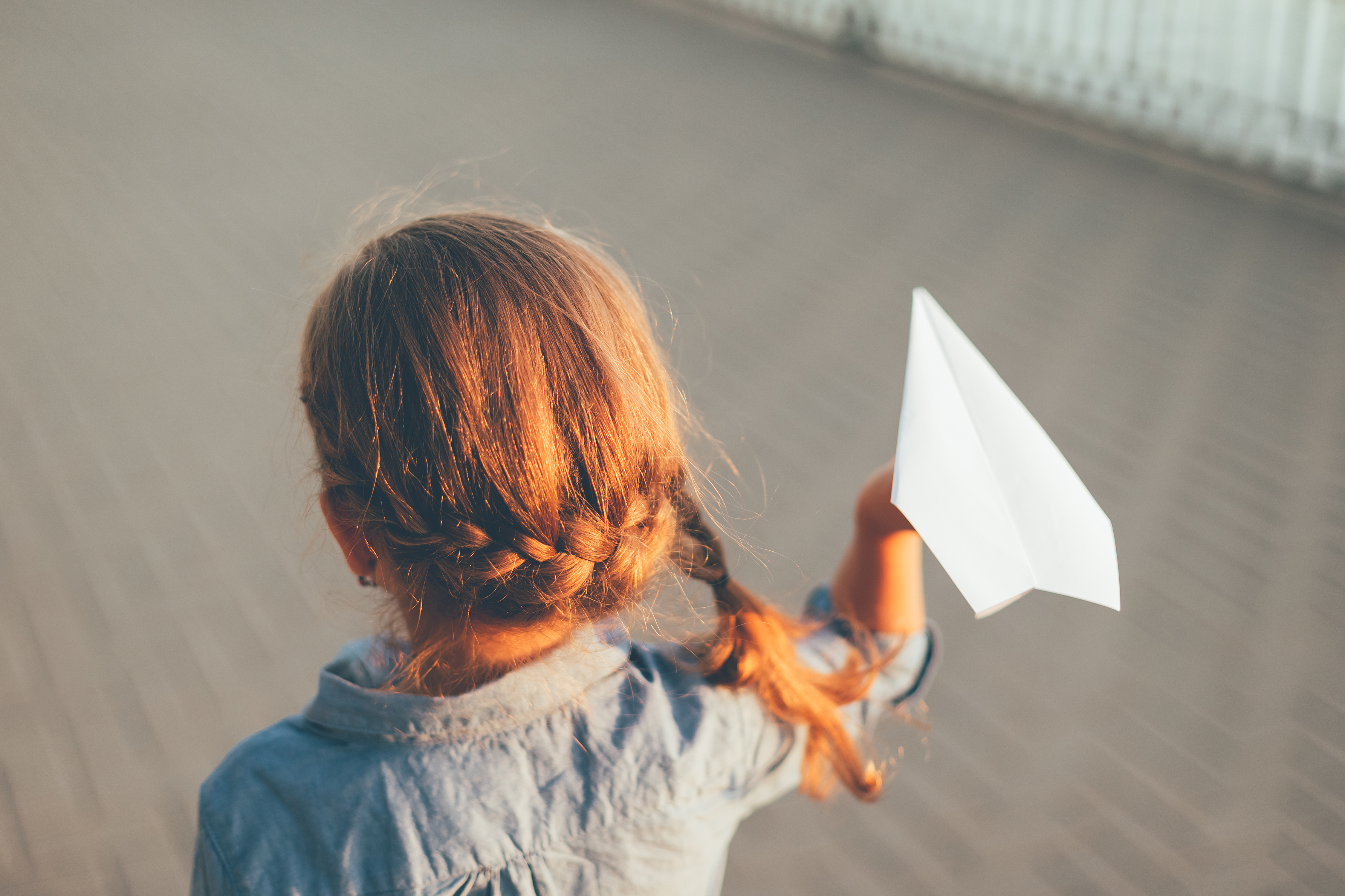 Close-up back view of child playing with toy paper airplane in the park, on gray pavage near lake in denim dress. Smiling girl looking in the future. Kid holding paper plane with arm.